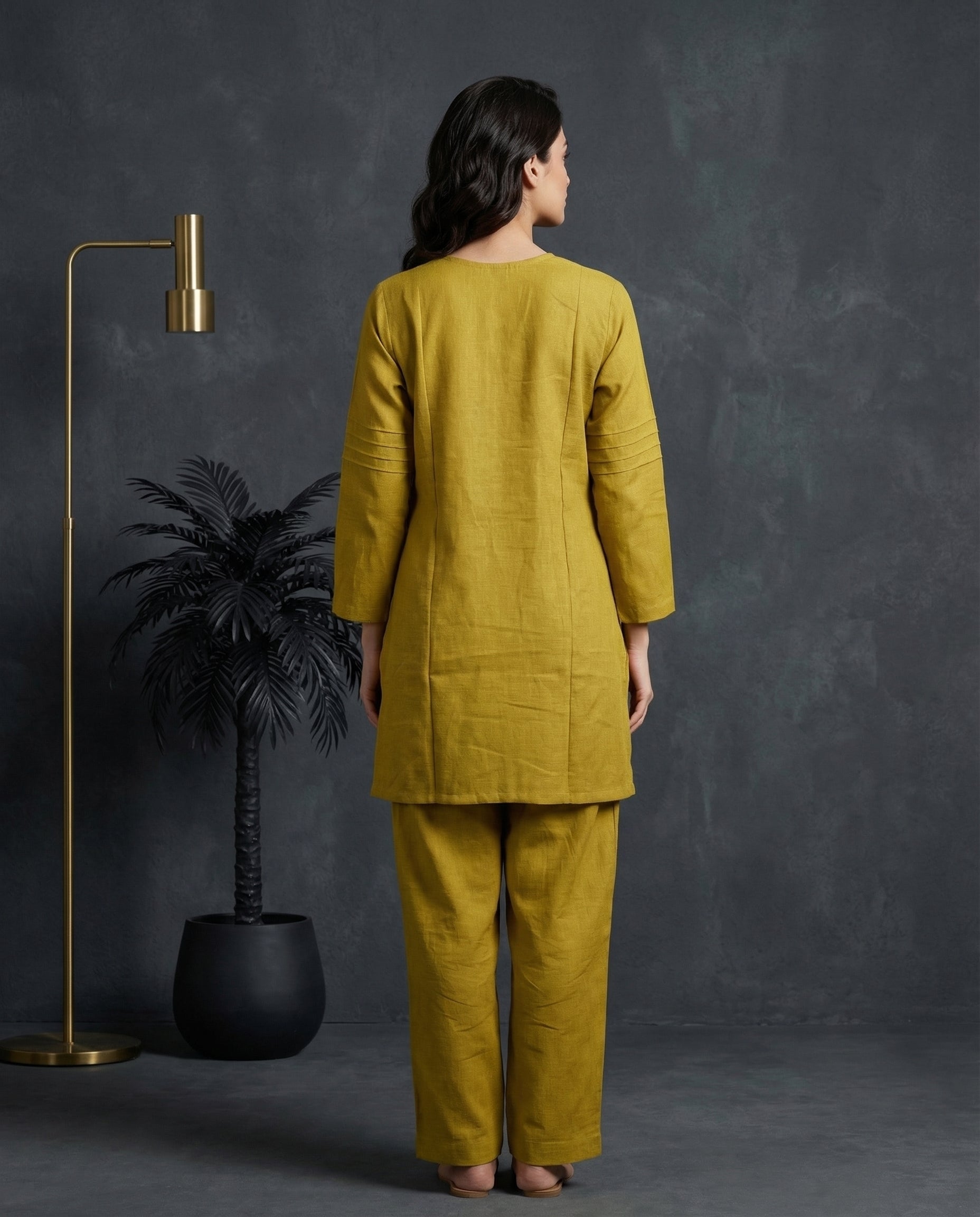 Woman wearing a mustard yellow outfit standing against a dark gray wall with a plant and lamp in the background.